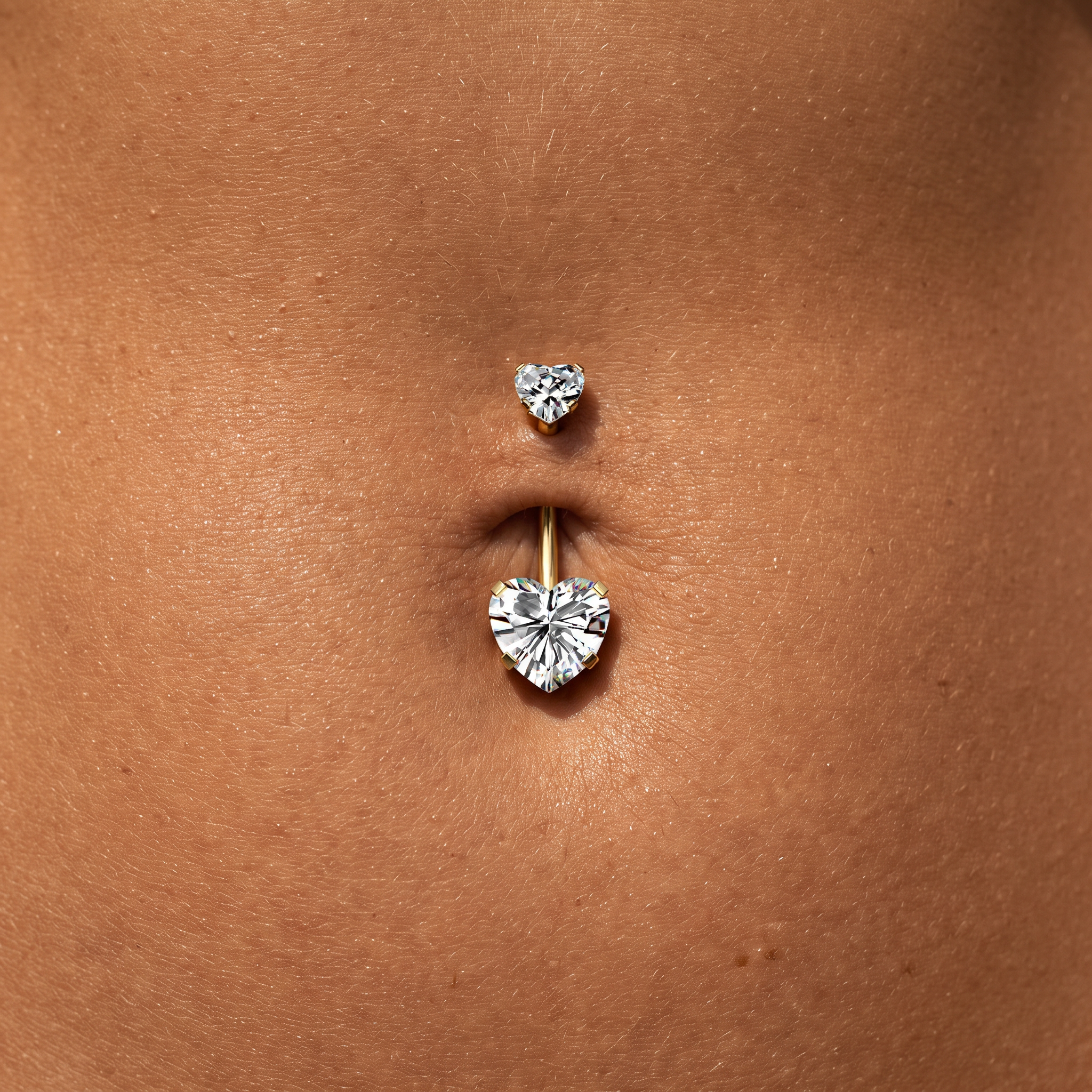 A close-up of a belly button piercing with a gold barbell featuring two heart-shaped clear gemstones, shown on a person's skin.