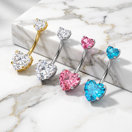 Four heart-shaped barbell belly button piercings arranged diagonally on a white marble slab. The jewelry features heart-shaped and round-cut clear, silver, pink, and blue stones in gold, silver, and white metal settings.