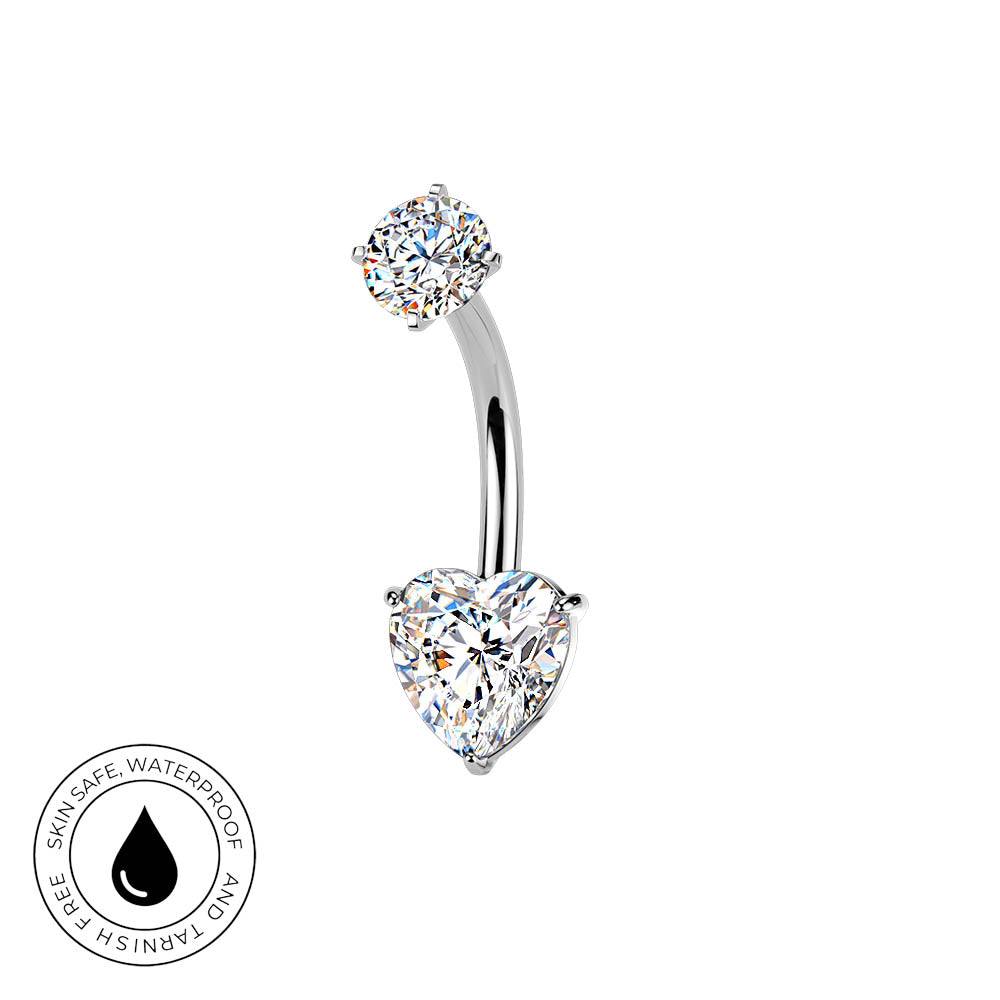A curved barbell belly button ring with a heart-shaped clear gemstone on the bottom and a round clear gemstone on the top, set in silver-toned metal. A circular badge in the bottom-left corner reads 'SKIN SAFE WATERPROOF AND TARNISH FREE' with a black droplet symbol.