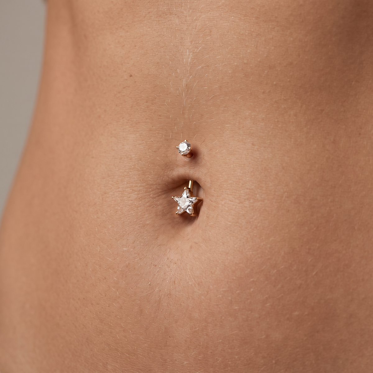 A close-up of a person's navel area showing two small belly button piercings: a simple round stud above and a gold star-shaped stud below.