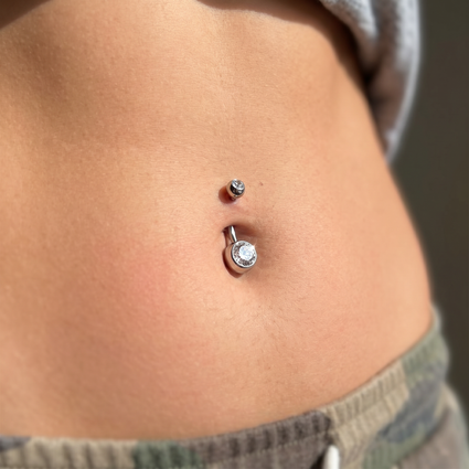 Close-up of a person's lower torso showing a belly button piercing set with a small silver stud above and a larger silver barbell with a clear rhinestone center below.
