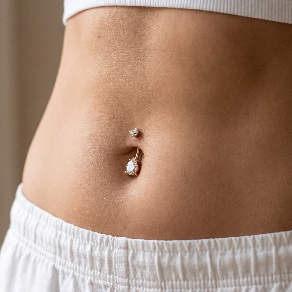 A close-up of a person's midsection showing a belly button jewelry set with a small silver stud and a larger gold-toned ring with a clear teardrop gemstone, worn with white clothing.