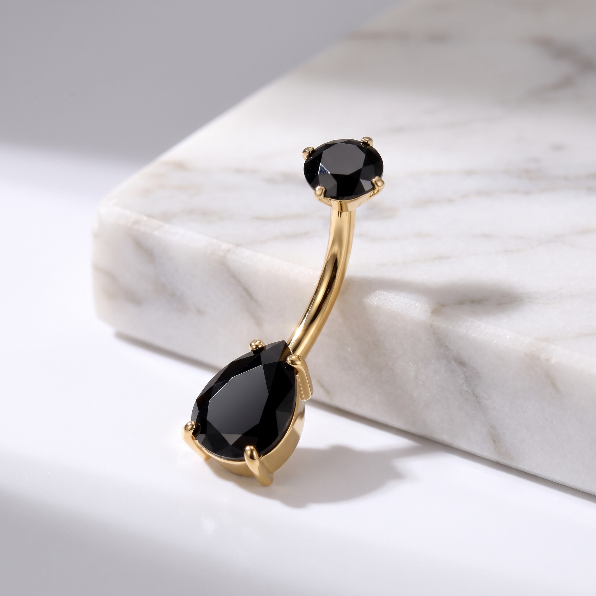 Gold curved barbell navel piercing jewelry with two black faceted gemstones, displayed on a white marble block.