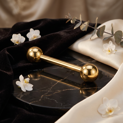 A polished gold-tone metal barbell with spherical ends rests on a black marble tray, surrounded by draped black velvet fabric, cream satin, white flowers, and a sprig of eucalyptus.
