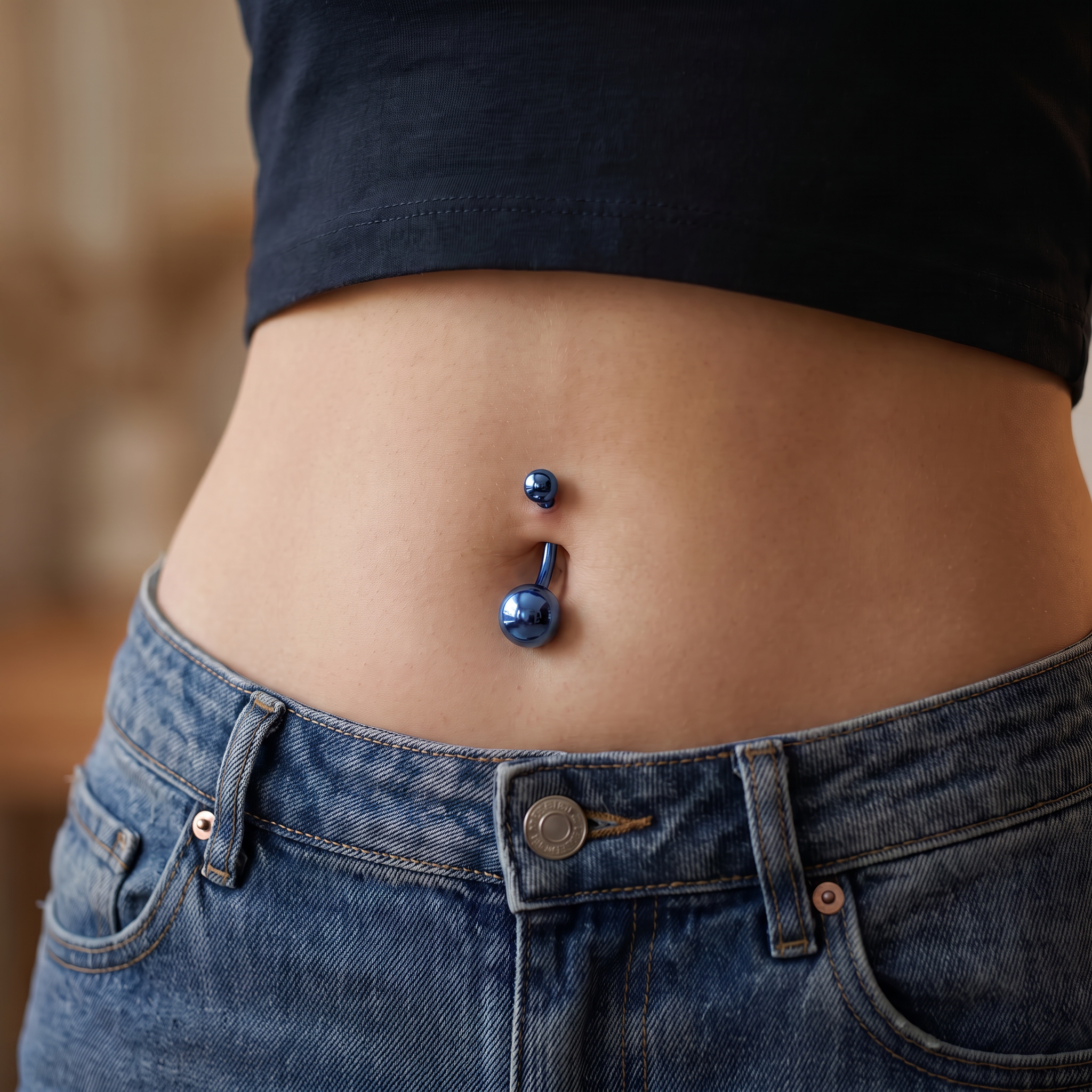 Close-up of a person's lower torso wearing blue jeans and a black crop top, showing a blue metallic navel piercing with two small blue studs.