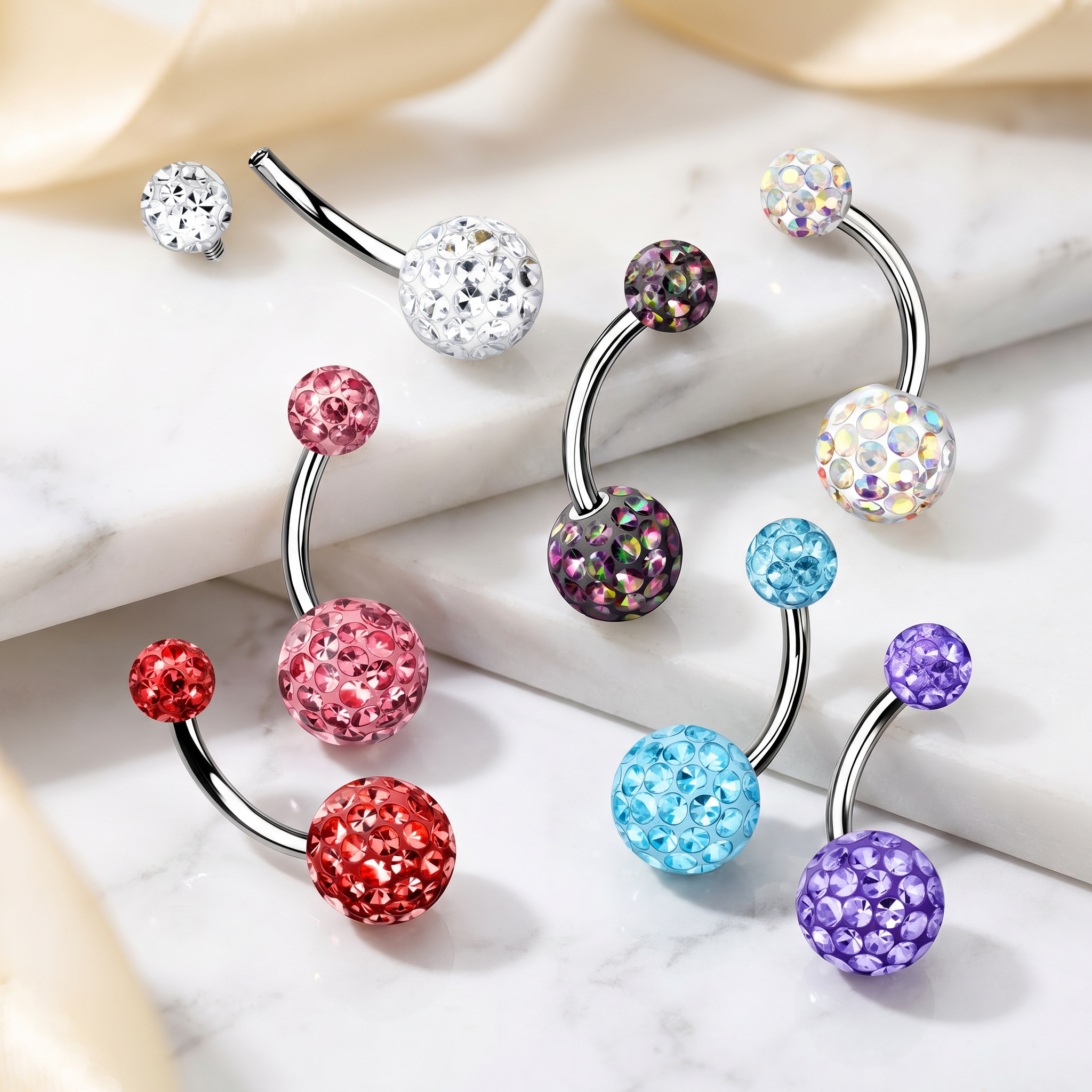 Four curved barbell belly button piercings with sparkling rhinestone-covered balls in assorted colors, arranged on a light marble surface with soft cream fabric in the background.