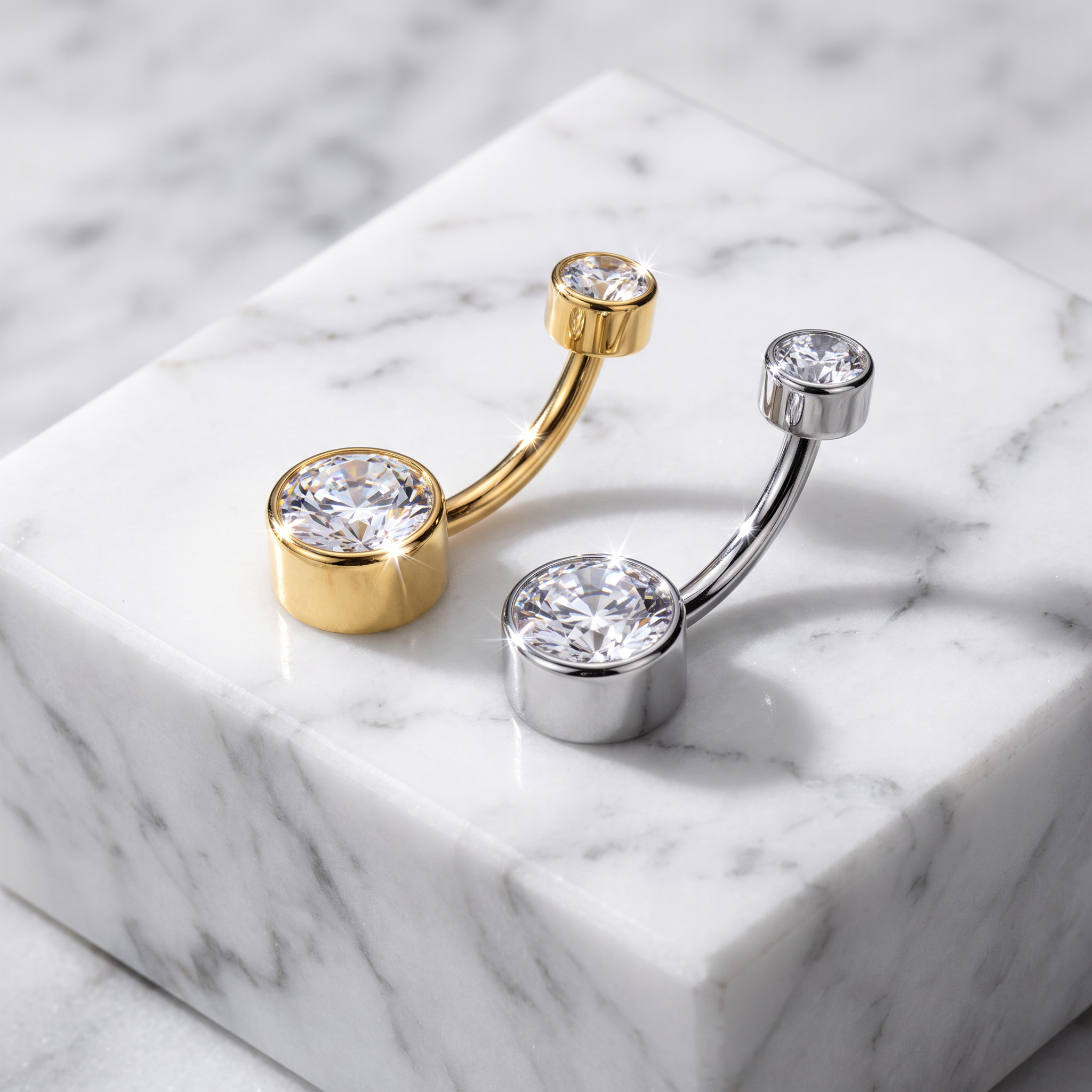 Two curved barbell belly button piercings displayed on a white marble block. One is gold-toned with a large clear gemstone, and the other is silver-toned with a matching large clear gemstone.