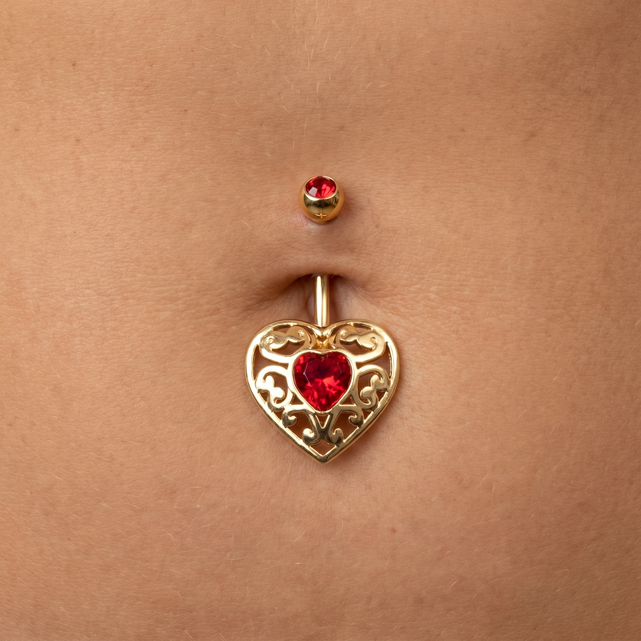 Gold belly button ring with an ornate filigree heart pendant featuring a red heart-shaped gemstone, shown worn on a person's navel.