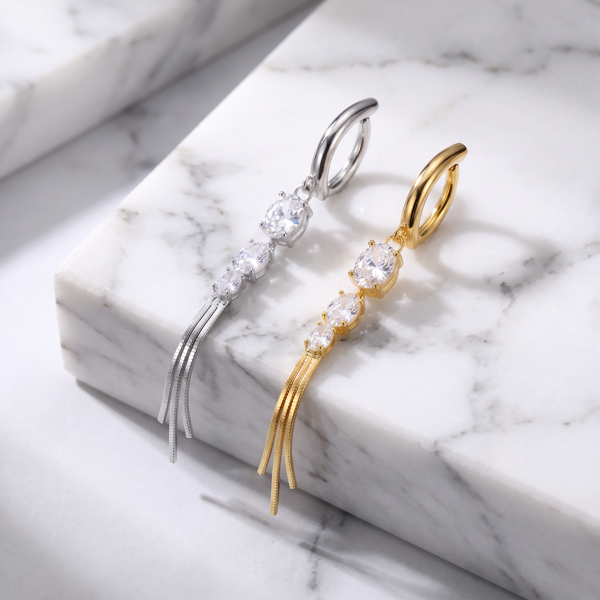 Two drop earrings displayed on a white marble block. One earring is silver-toned with a large clear stone, three smaller clear stones, and three dangling thin strands.