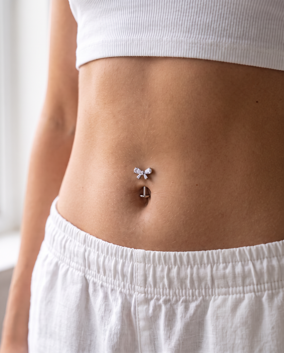Close-up of a person's midsection wearing a small silver belly button ring with a bow and anchor design, paired with a white ribbed crop top and white shorts.