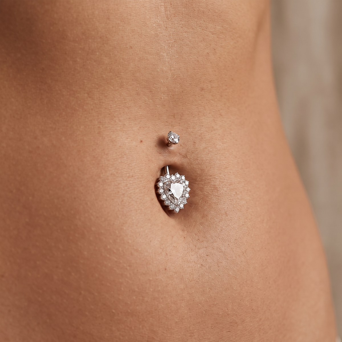 A close-up of a person's lower torso showing a belly button with two silver-toned navel piercings: a small round stud above and a larger heart-shaped halo stud below. The heart stud features a clear teardrop gemstone surrounded by small clear stones.