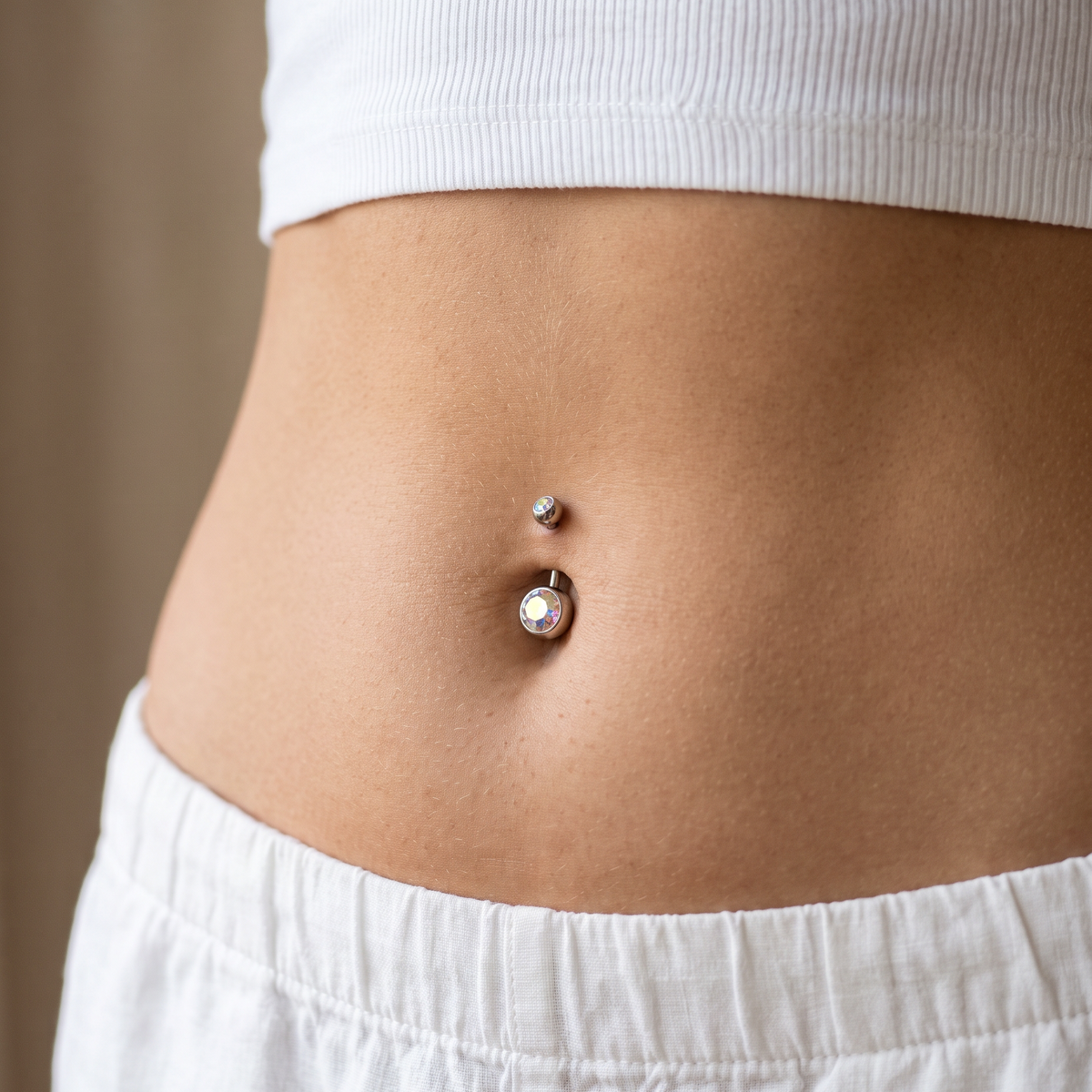A close-up of a person's lower torso showing a double-piece belly button jewelry set, featuring a small silver stud and a larger charm with a clear rhinestone, worn on natural skin.