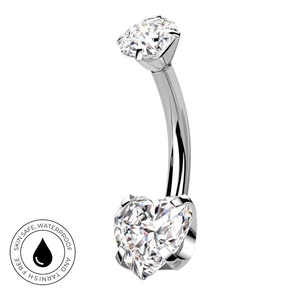 A curved barbell belly button ring with a large heart-shaped clear gemstone on one end and a round clear gemstone on the other, shown on a white background with a small circular skin-safe waterproof badge in the lower left.