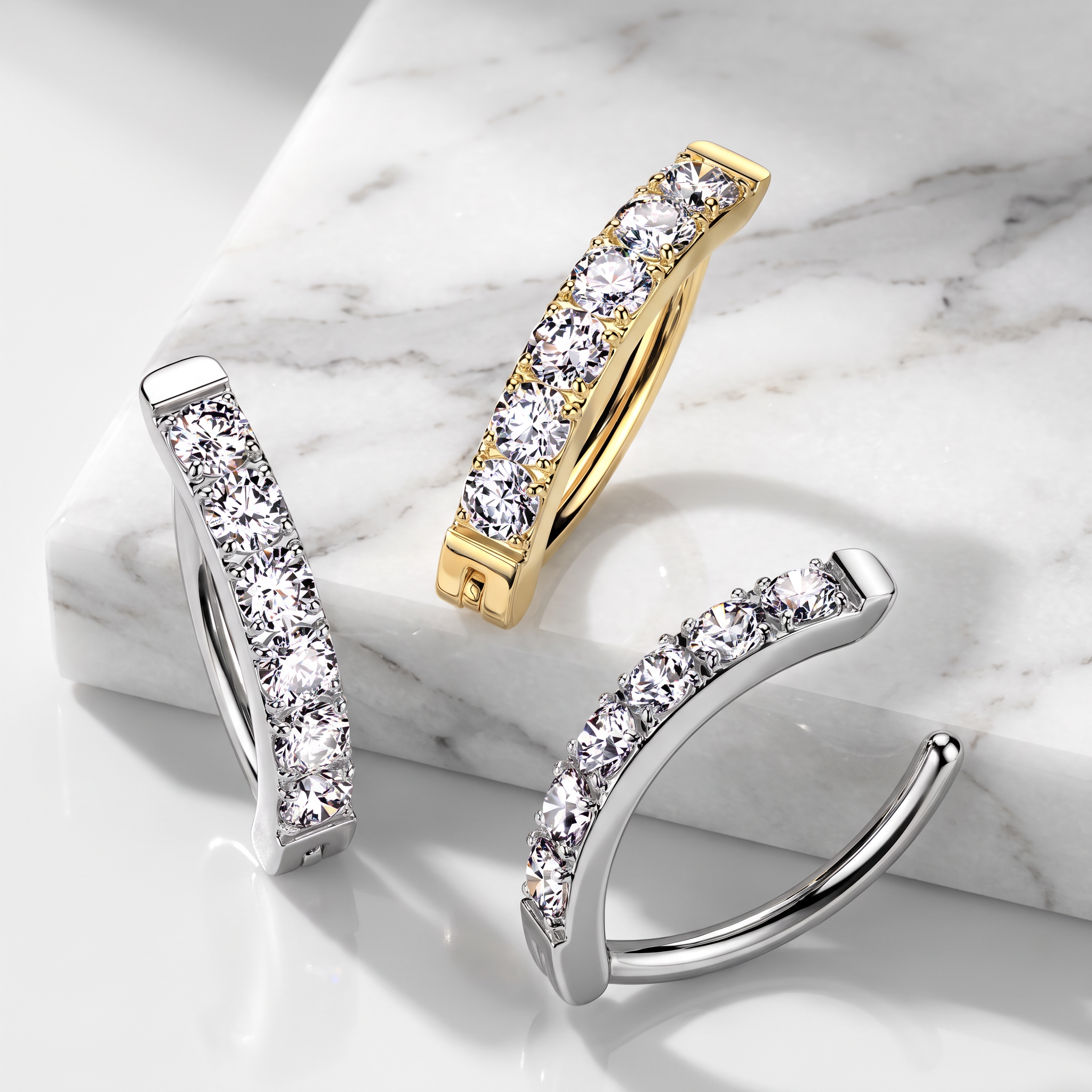 Three curved huggie hoop earrings with rows of clear pavé stones, shown in silver and yellow gold finishes on a white marble block and white surface.
