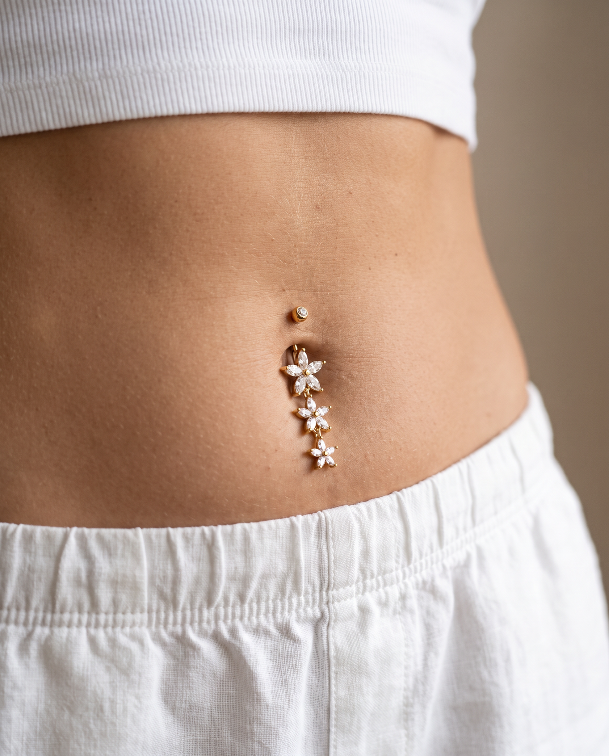 A close-up of a person's lower torso wearing a rose gold belly button ring with three dangling flower charms and a small gold bead, styled with white ribbed crop top and white pants.