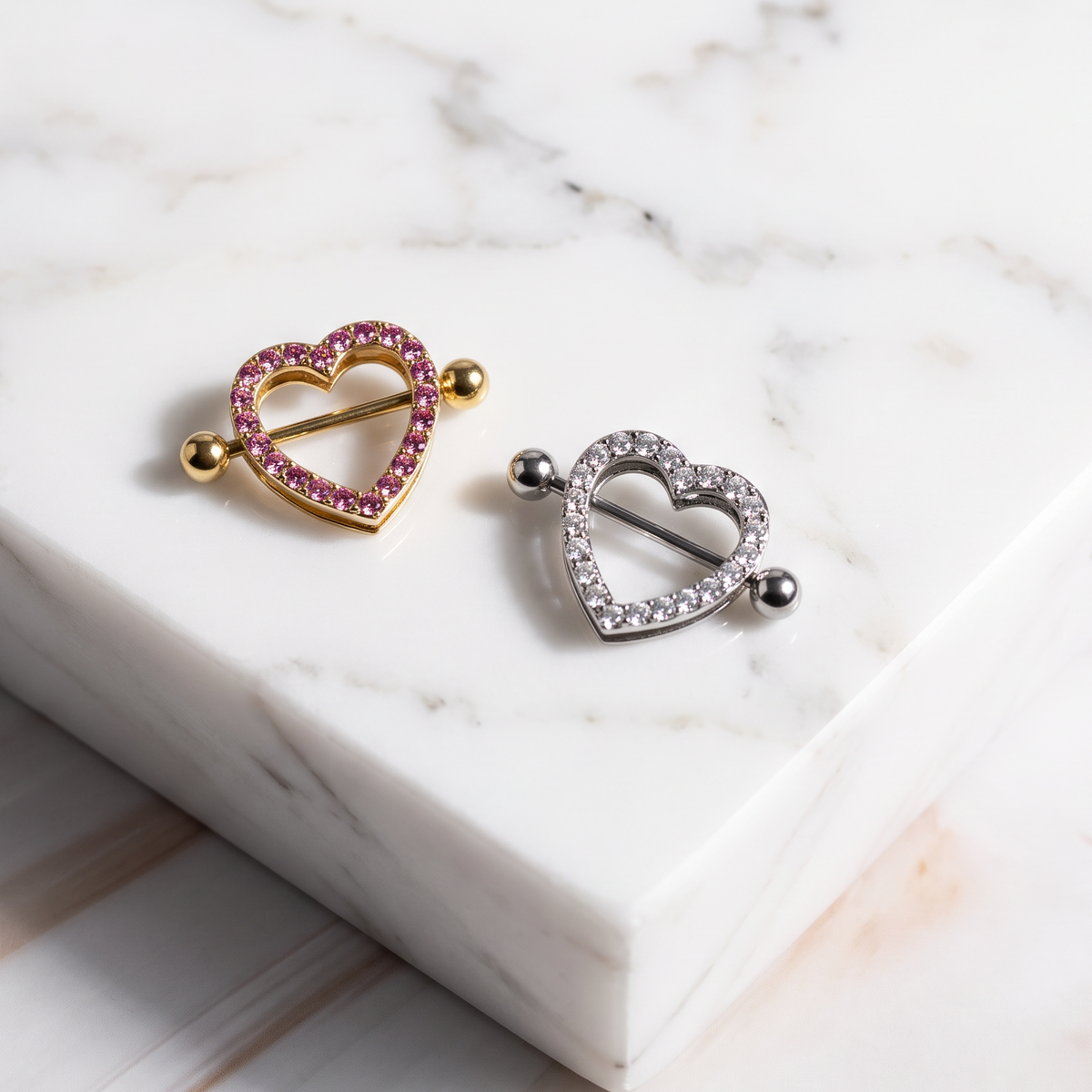 Two heart-shaped nipple shields displayed on a white marble block. One earring is gold with pink stones, and the other is silver with clear stones.