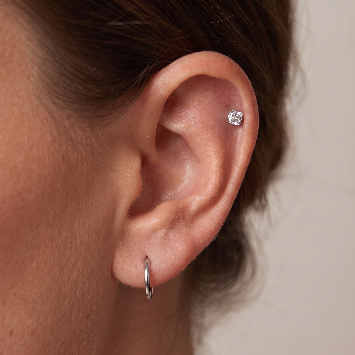 Close-up of a person's ear showing two silver earrings: a small hoop earring in the lobe and a round stud earring in the cartilage, against a plain light beige background.