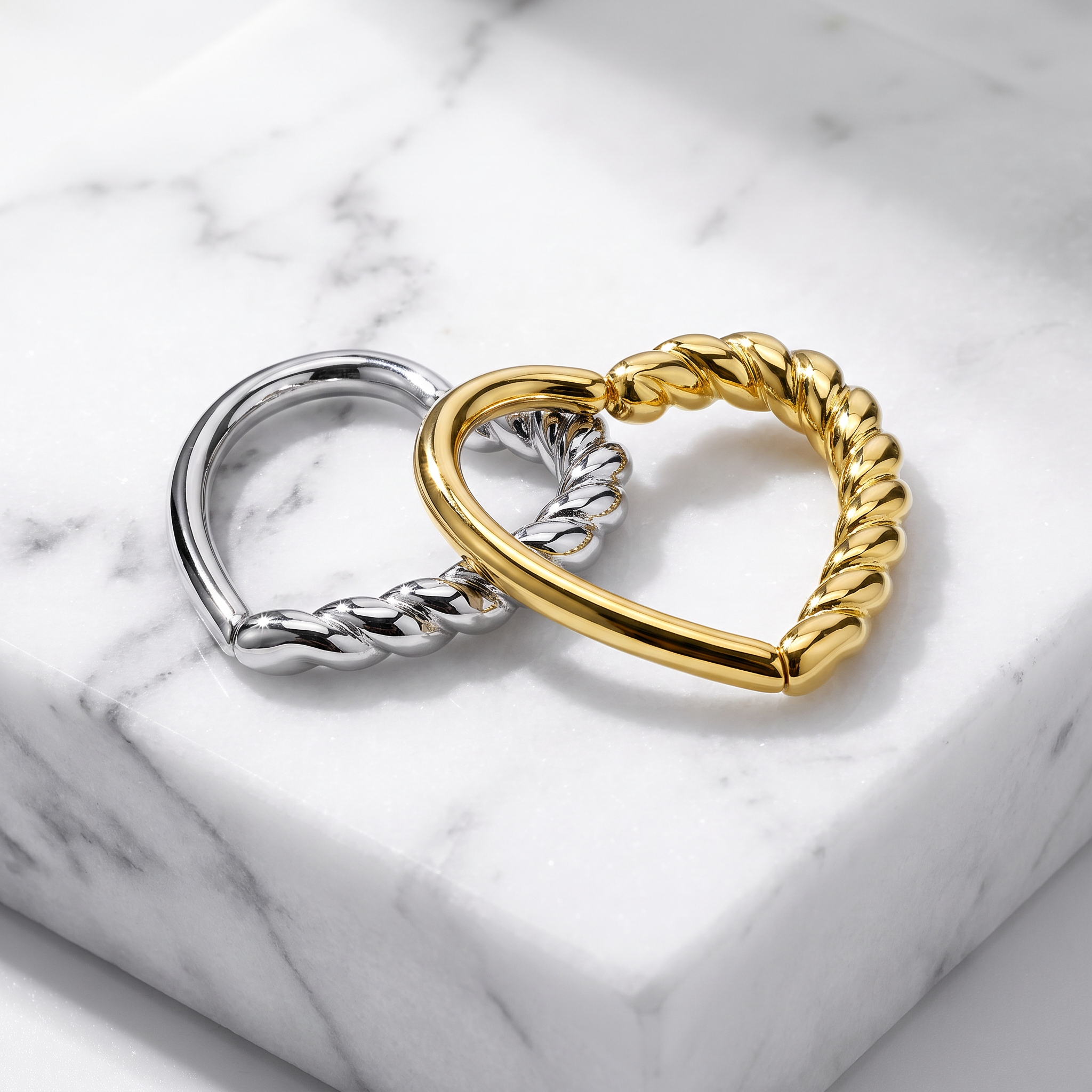 Two twisted rope-style metal rings, one polished gold and one polished silver, displayed on a white marble block.