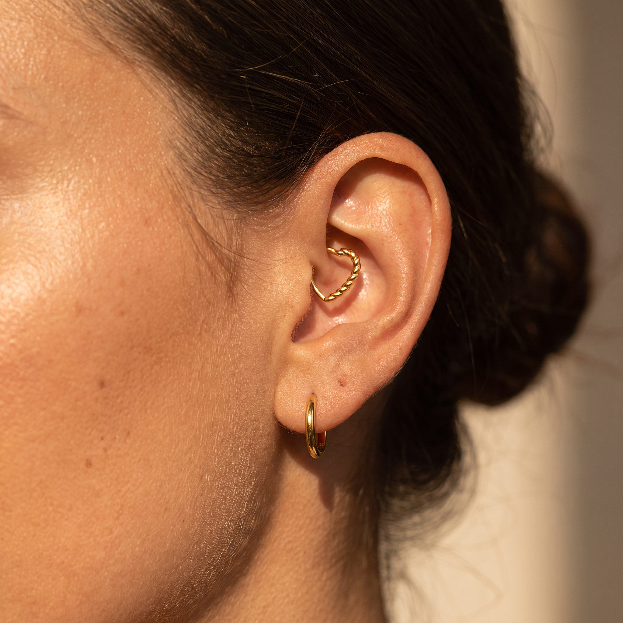 A close-up of a person's ear showcasing a gold ear cuff with a twisted heart design and a small gold hoop earring, set against a warm neutral background.