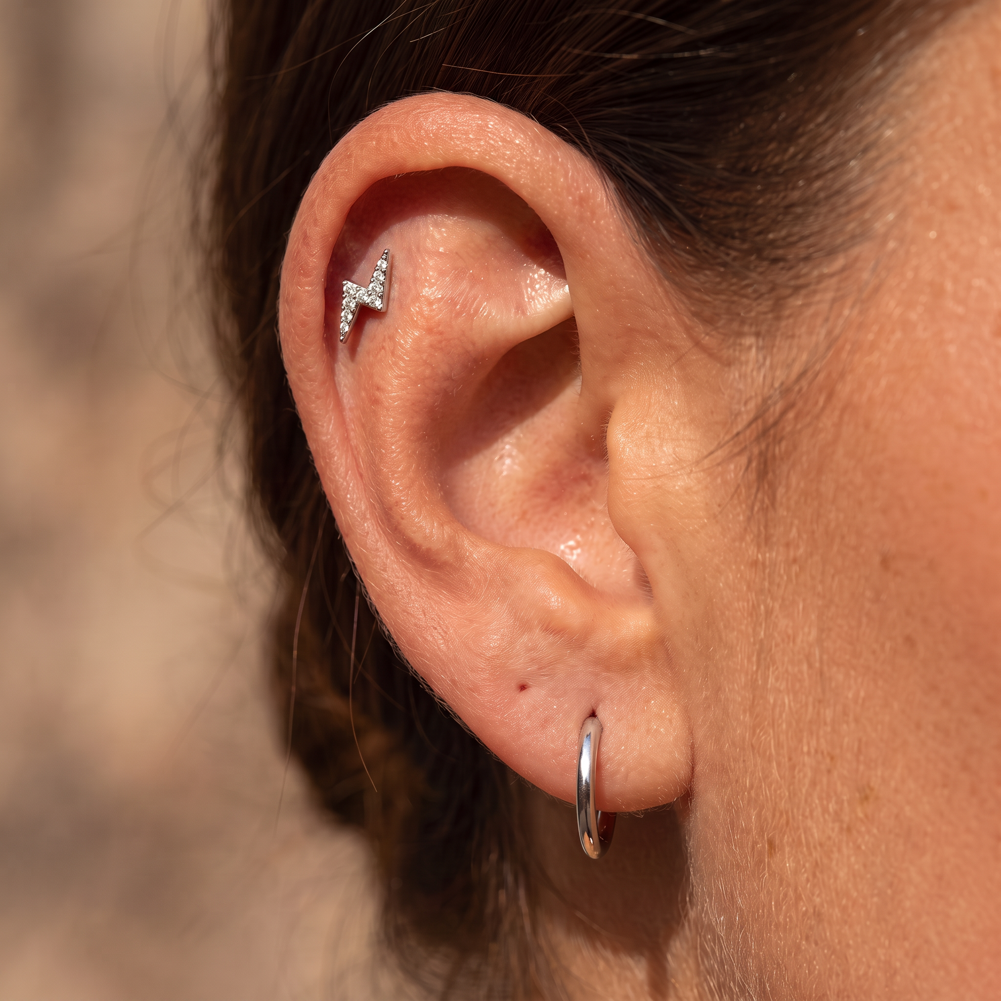 Close-up of a person's ear showcasing a silver lightning bolt stud earring in the helix and a simple silver hoop earring in the lobe, set against a blurred warm beige background.