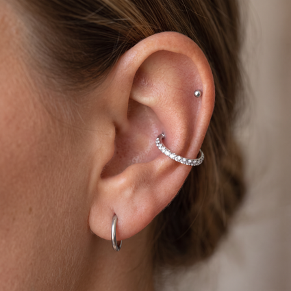 Close-up of a person's ear showcasing silver body jewelry, including a silver hoop in the lobe, a silver cartilage hoop adorned with small clear stones, and a silver dome stud in the rib cage.