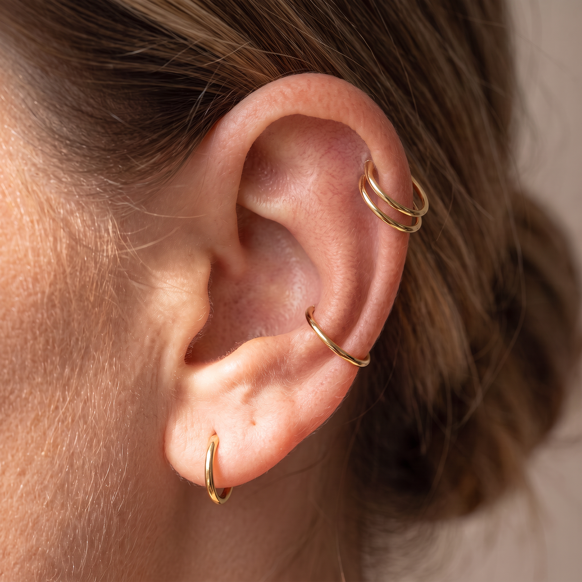 A close-up of a person's ear adorned with three delicate gold ear piercings: a small hoop in the lobe, a curved barbell cartilage piercing, and a double-ring hoop in the helix.