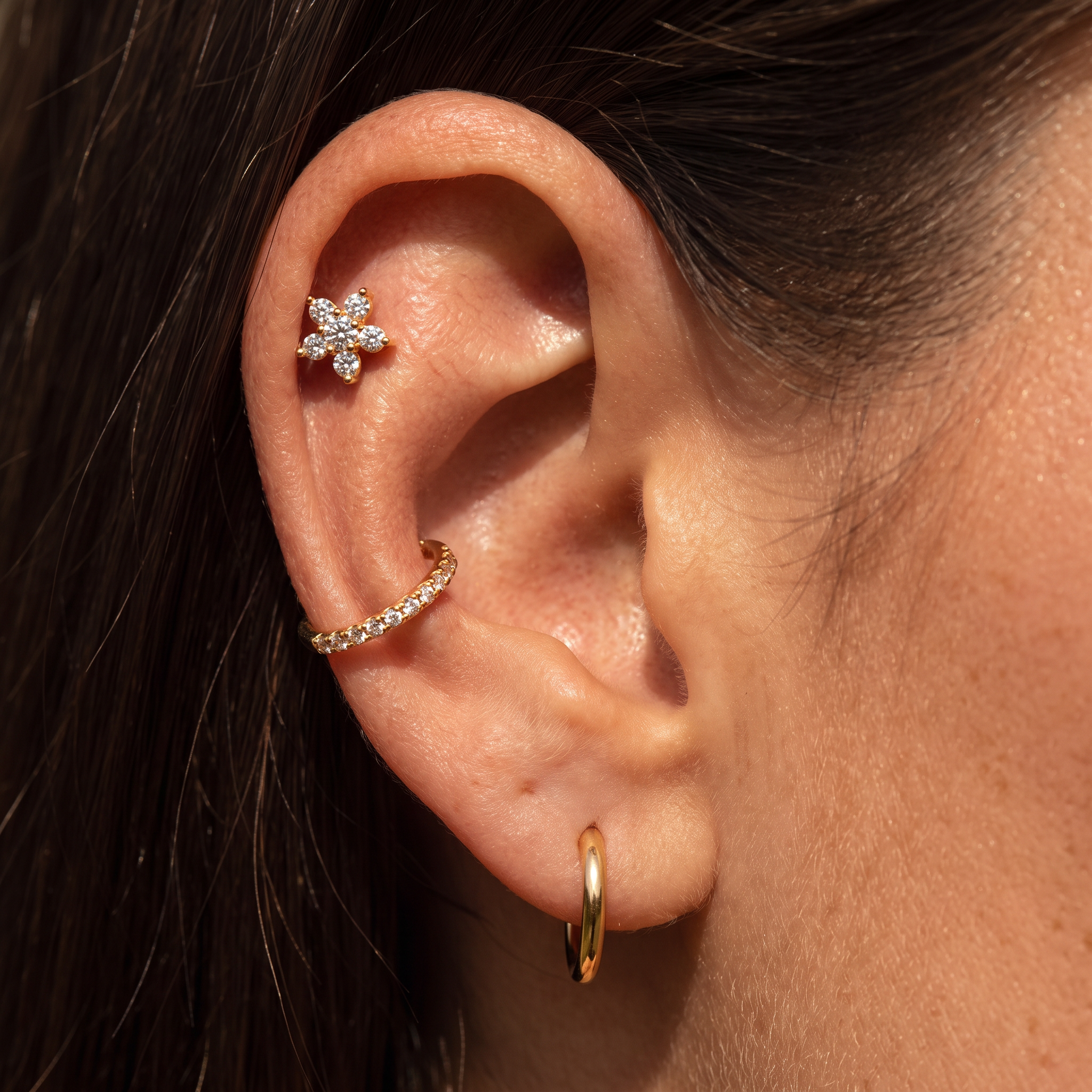 Close-up of a person's ear showcasing three gold ear piercings: a small flower-shaped stud, a curved hoop set with clear stones, and a simple gold hoop earring.