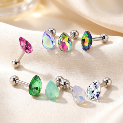 Six silver-toned barbell stud earrings with teardrop-shaped faceted gemstones in pink, iridescent blue/purple, iridescent green/blue, iridescent blue/purple, green, and pale blue, arranged on a beige satin fabric background.