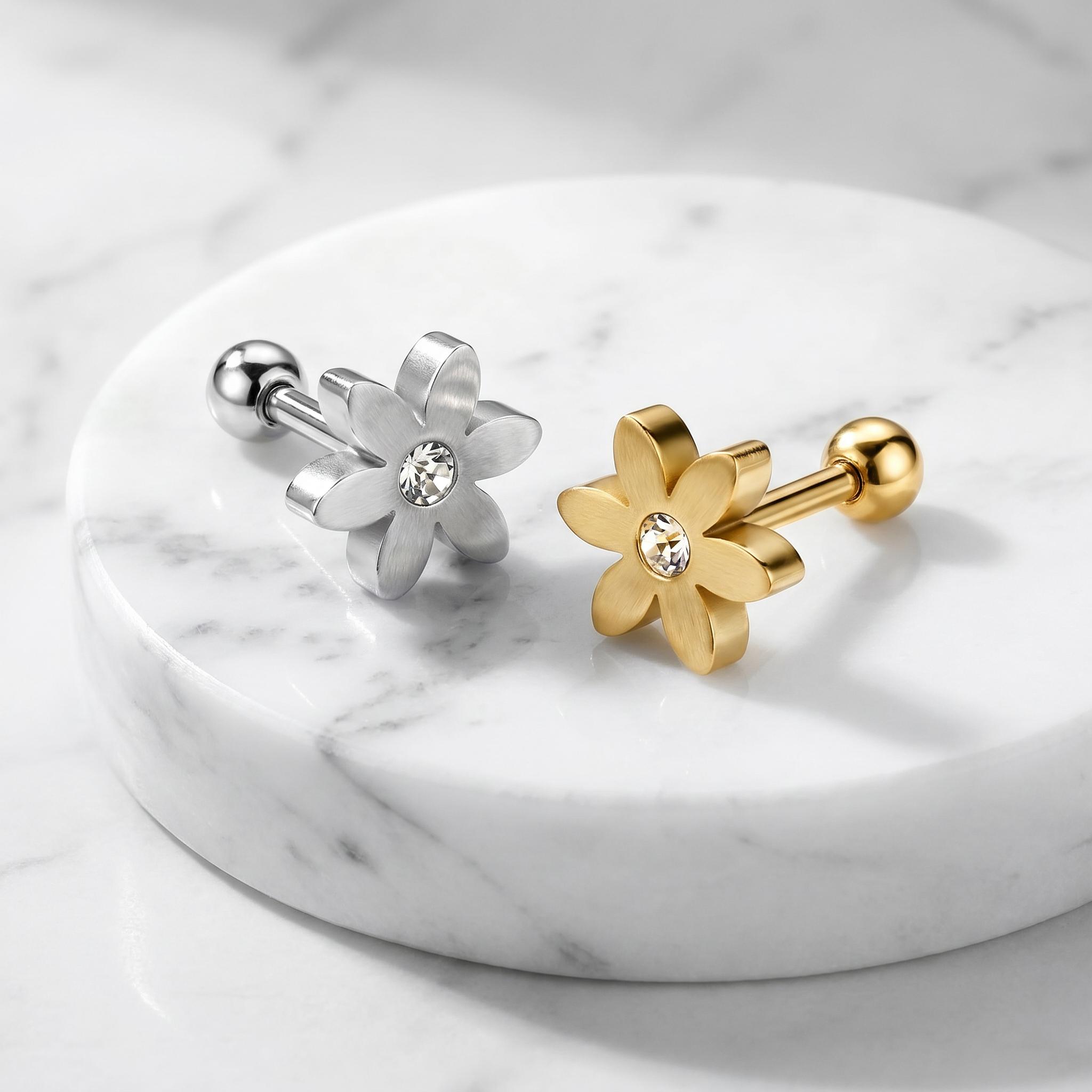 Two flower-shaped stud earrings displayed on a round white marble tray. One earring is silver-toned with a clear crystal center, and the other is gold-toned with a clear crystal center.