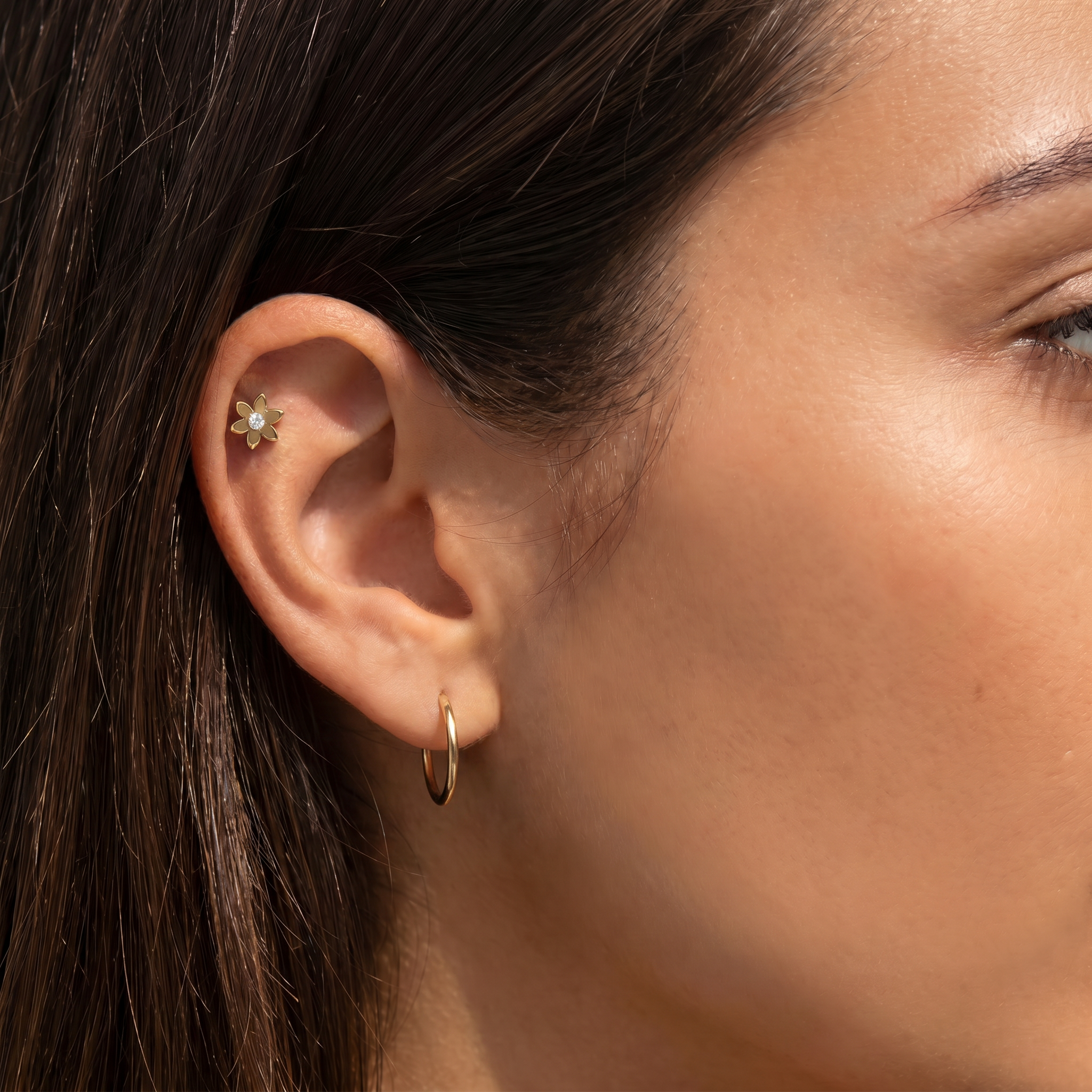 Close-up of a woman's ear showing two gold earrings: a small flower-shaped stud in the helix and a delicate hoop in the lobe.