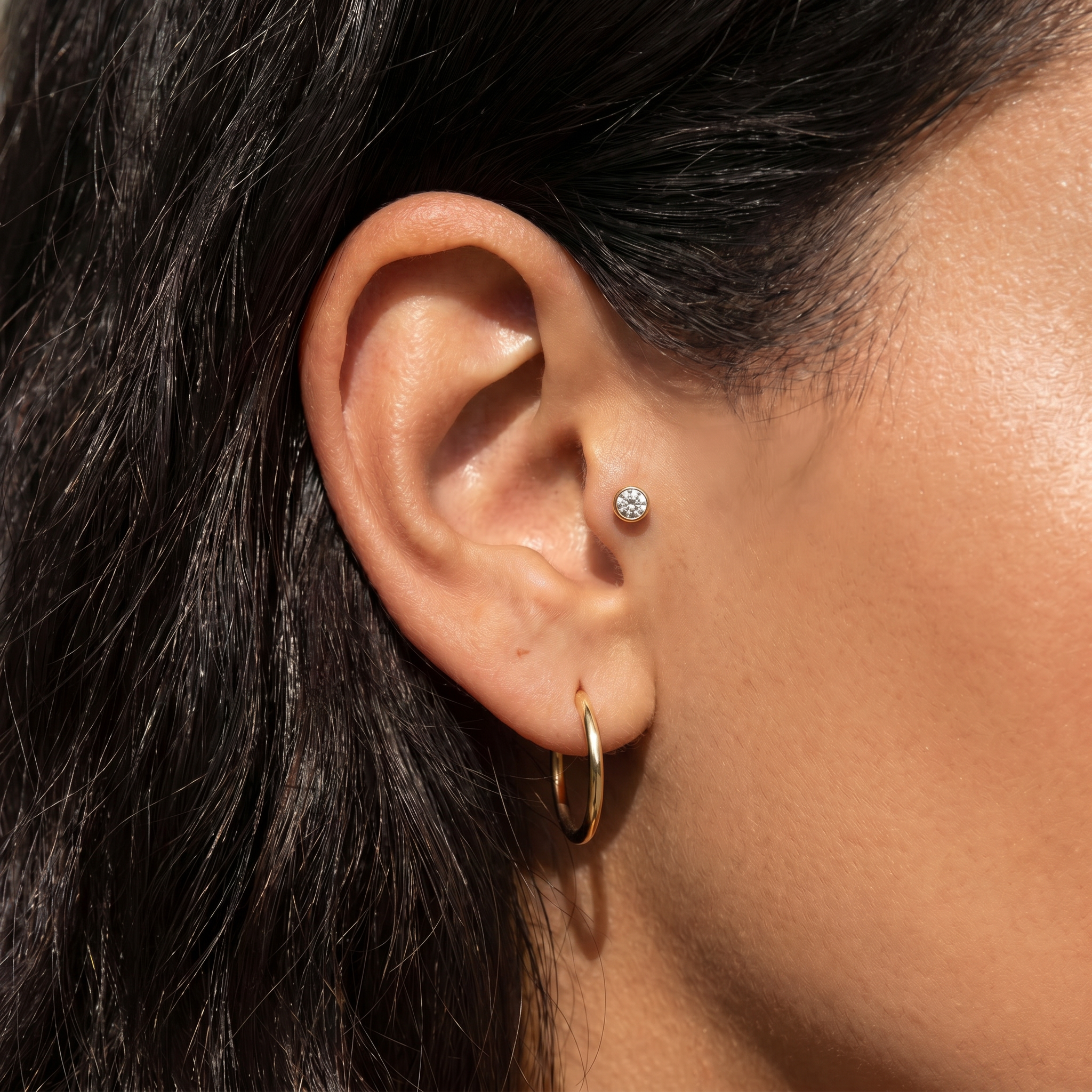 Close-up of a person's ear wearing two earrings: a small round clear gemstone stud on the helix and a gold hoop earring on the lobe.