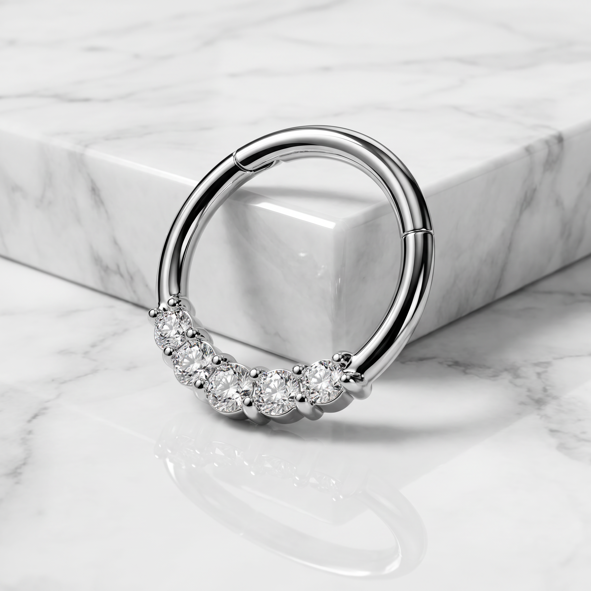 A silver-toned hoop ring with a curved row of clear gemstones along the lower front, photographed on a white marble surface with a matching marble block behind it.