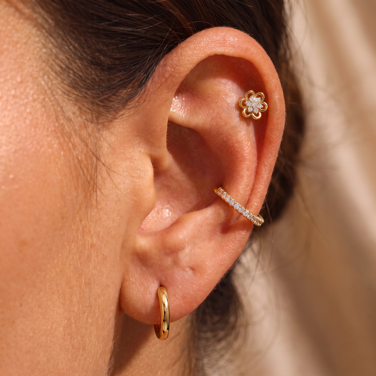 A close-up of a person's ear showcasing a gold ear stacker set with three pieces: a small flower-shaped stud, a curved ear cuff lined with clear stones, and a simple gold hoop earring.