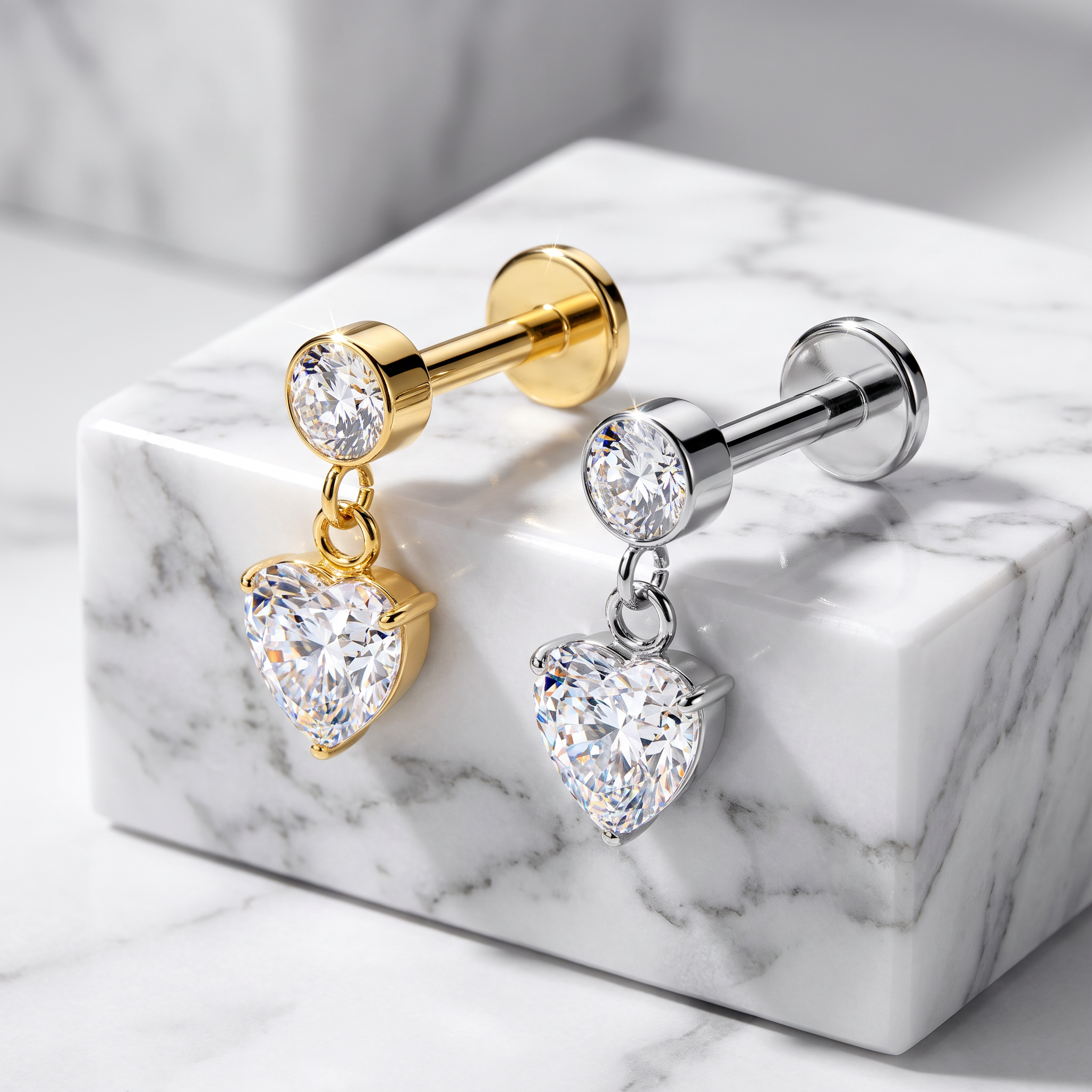 Two heart-shaped dangle earrings displayed on a white marble block. One earring features a gold-plated post and bezel with a clear heart-shaped gemstone, and the other features a silver-plated post and bezel with a matching clear heart-shaped gemstone.