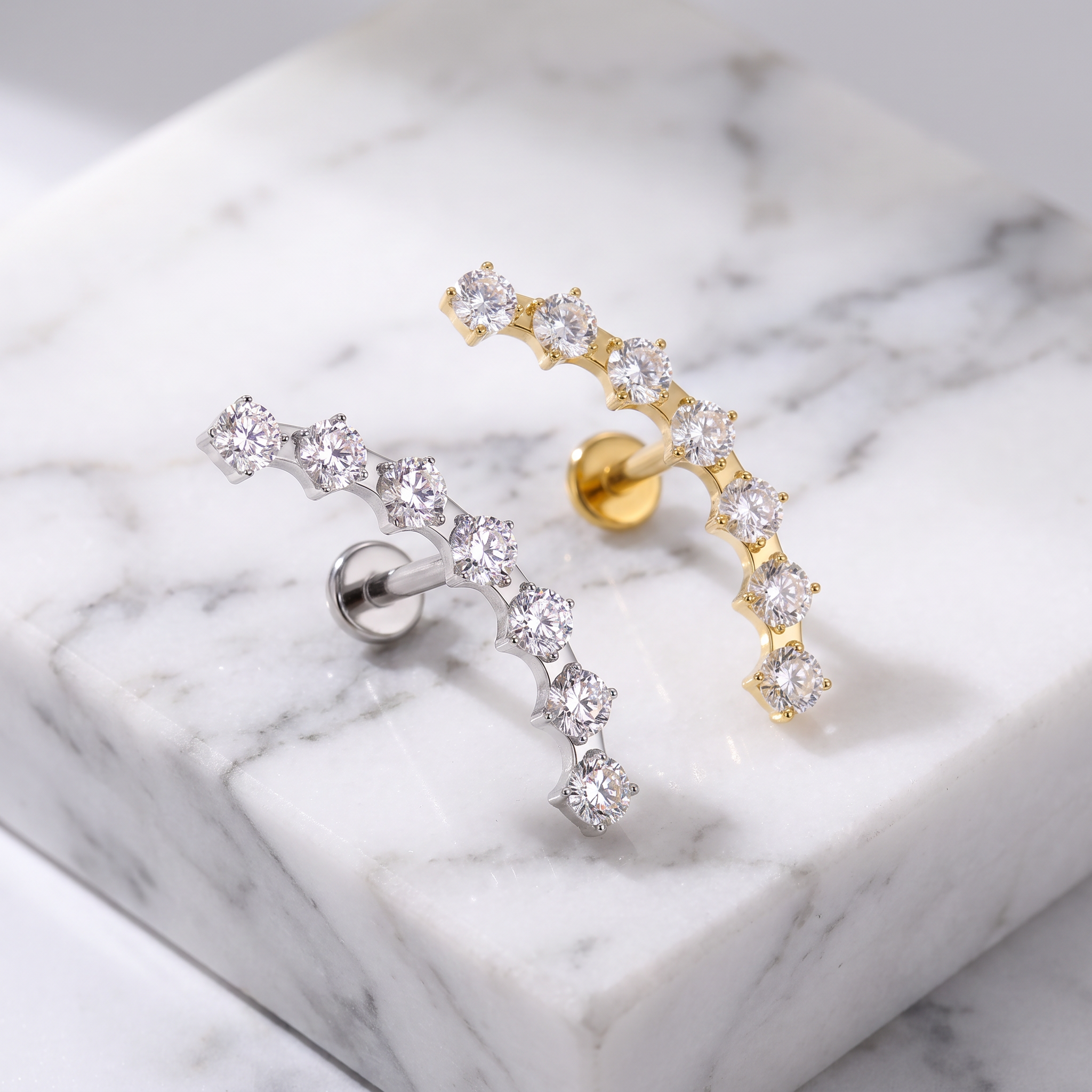 Two curved ear cuff earrings with clear cubic zirconia stones, one in yellow gold and one in silver-tone metal, displayed on a white marble block.