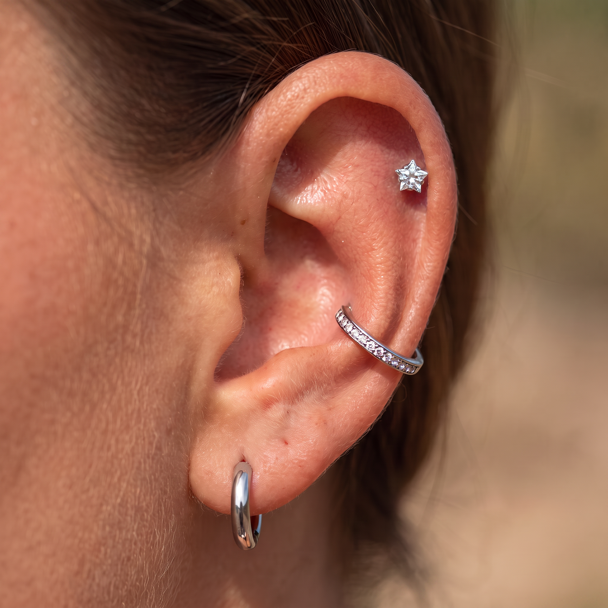 Close-up of a person's ear showcasing three silver ear piercings: a small star-shaped stud, a curved cartilage hoop set with clear stones, and a flat hoop earring.