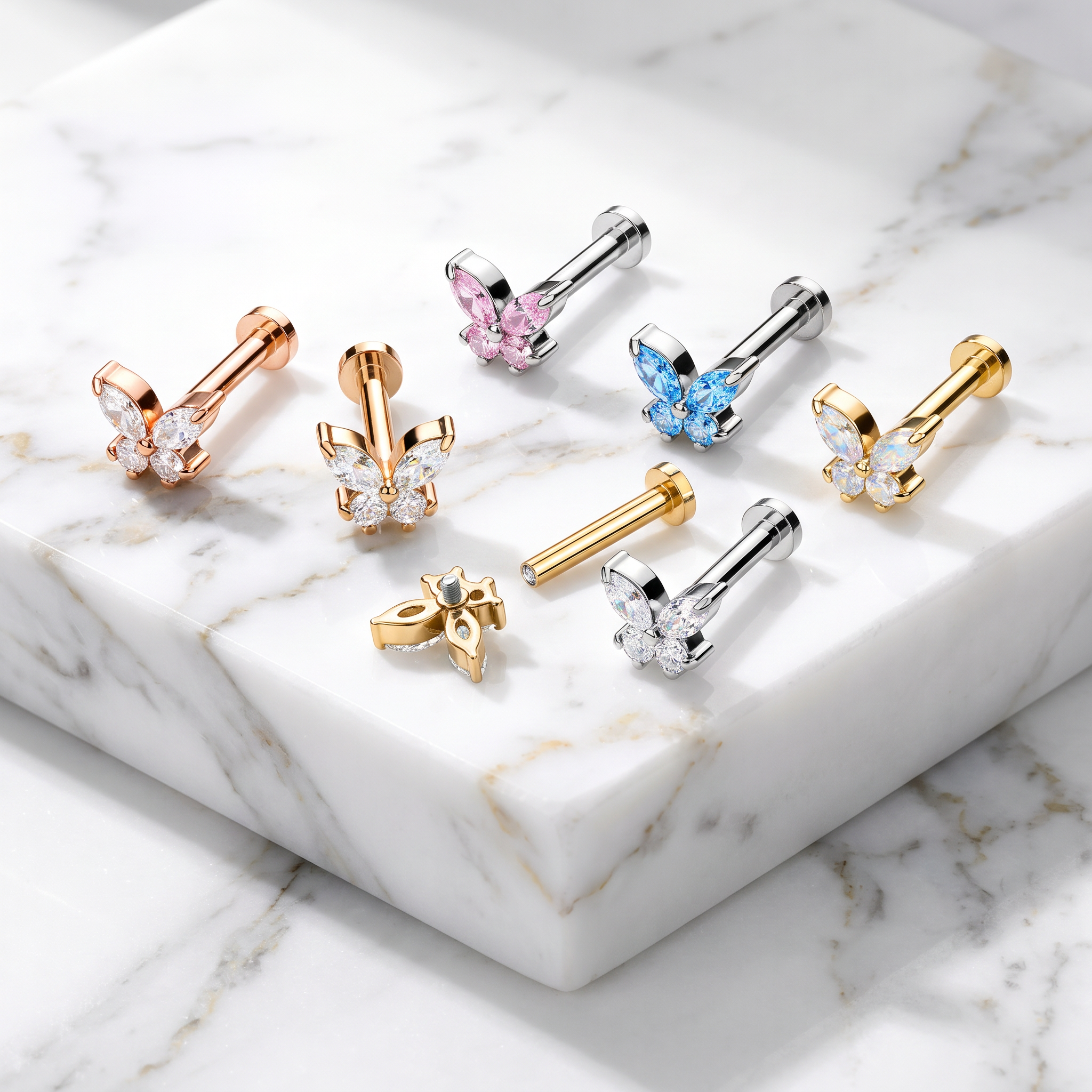 Six pairs of butterfly-shaped labret ear piercings in rose gold, yellow gold, and silver, some with clear rhinestones and others with pink, blue, and white rhinestones, arranged on a white marble block.