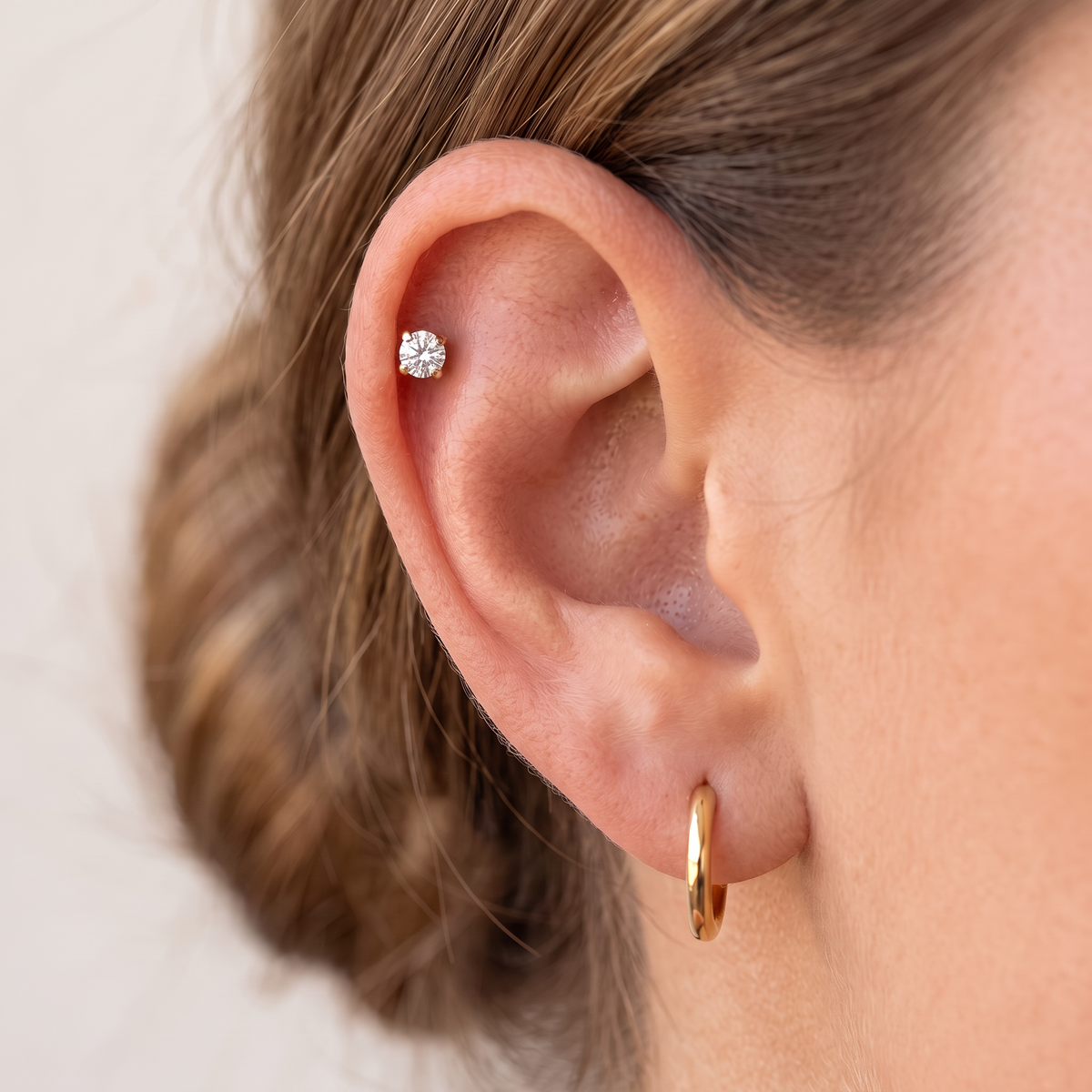 Close-up of a person's ear showcasing two earrings: a small round clear gemstone stud in the lobe and a delicate gold hoop in the cartilage.