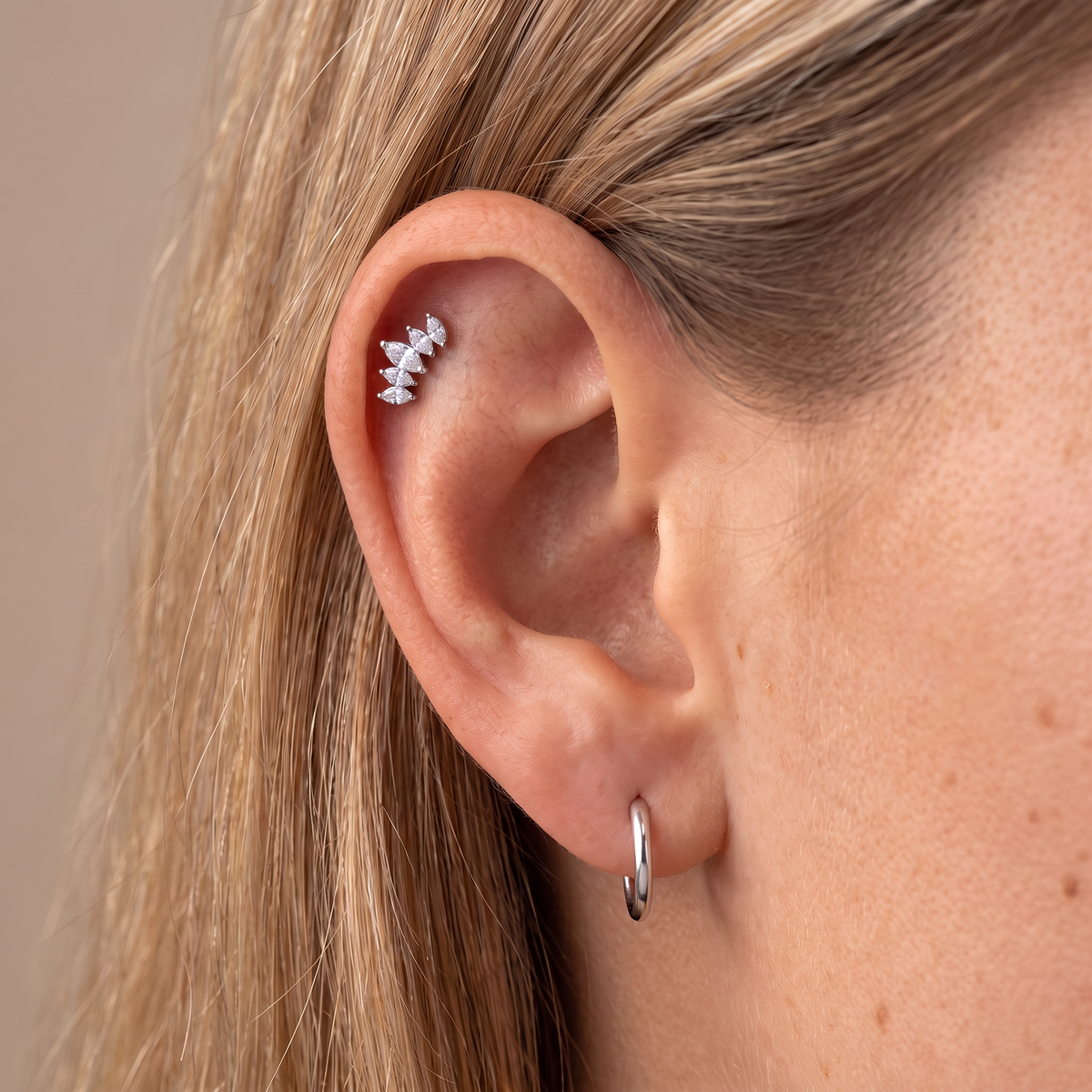 A close-up of a person's ear showing a small silver ear cuff with three clear gemstones and a silver hoop earring, against a soft beige background.