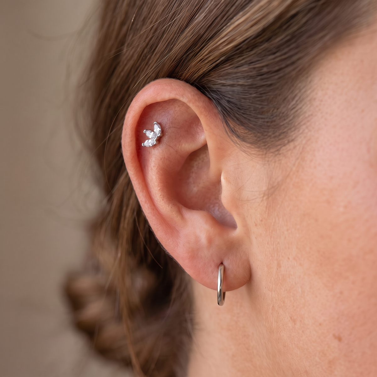 A close-up of a person's ear showcasing a small silver butterfly stud earring in the helix cartilage and a simple silver hoop in the lobe, against a soft beige background.