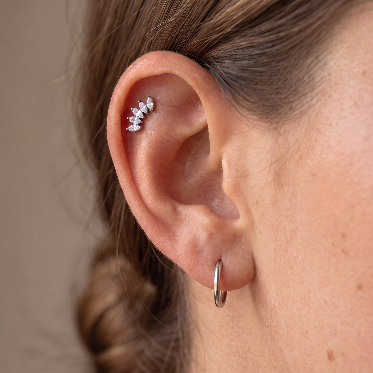 A close-up of a person's ear showcasing a silver leaf cluster ear cuff and a simple silver hoop earring against a neutral beige background.
