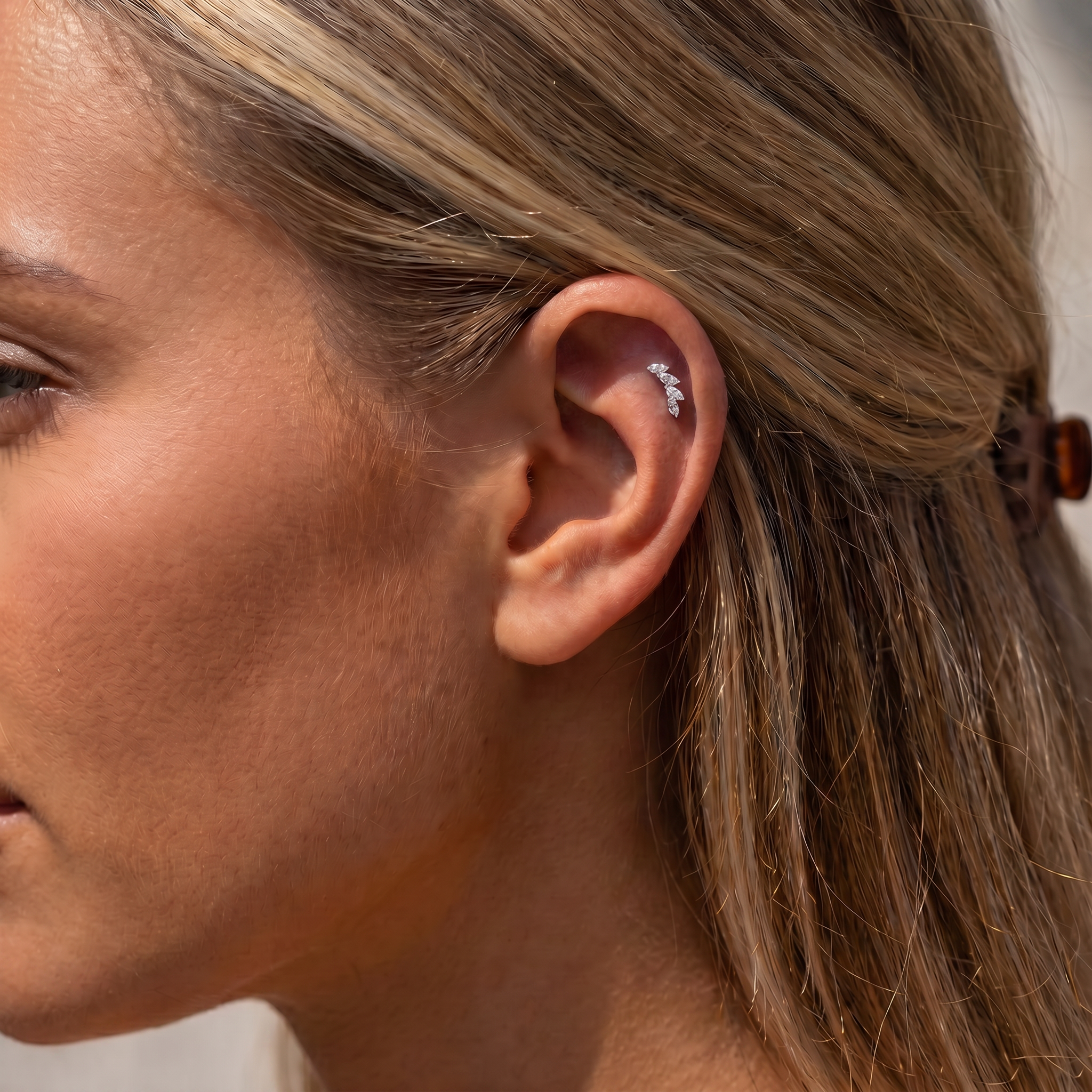 A close-up side profile of a woman's ear wearing a small silver cartilage piercing stud with clear stones.