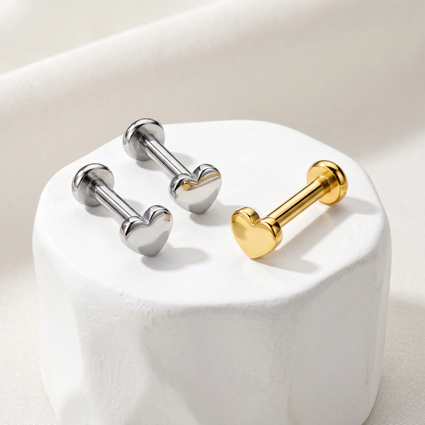 Three heart-shaped barbell stud earrings displayed on a white cylindrical pedestal. Two are silver-toned and one is gold-toned, set against a soft beige fabric background.