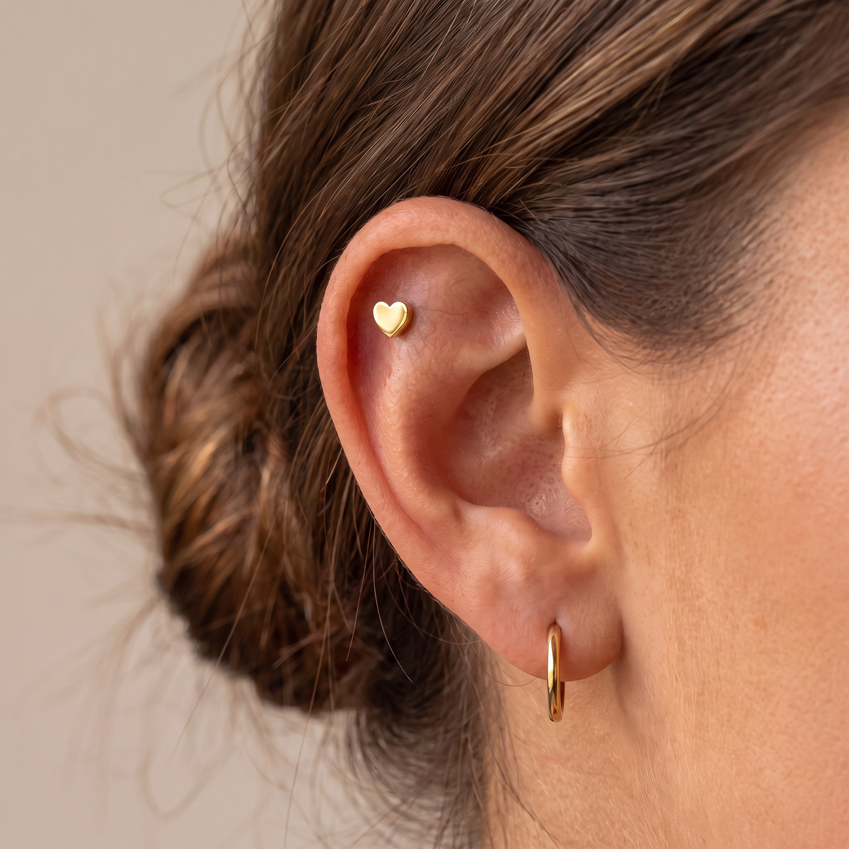 A close-up of a person's ear wearing a small gold heart stud earring in the helix and a thin gold hoop earring in the lobe, against a soft beige background.