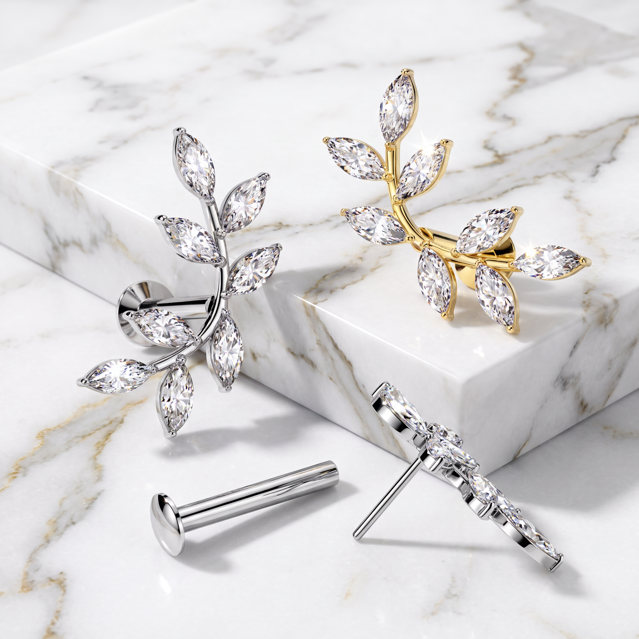 Two leaf-shaped stud earrings with marquise-cut clear stones, shown in silver and gold metal finishes on a white marble surface and block.