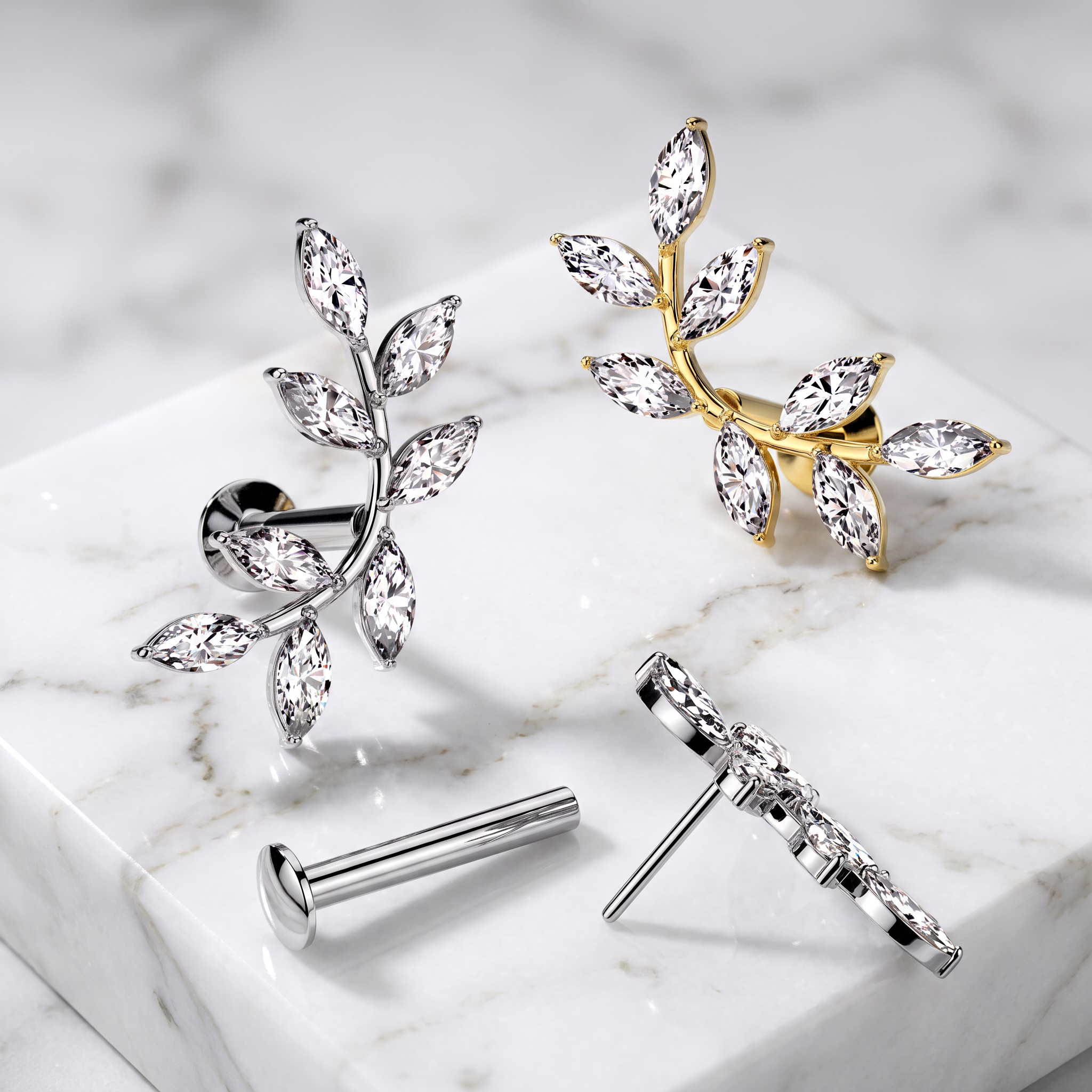 Two leaf-shaped stud earrings displayed on a white marble slab, one in silver and one in gold, with their posts shown separately below.