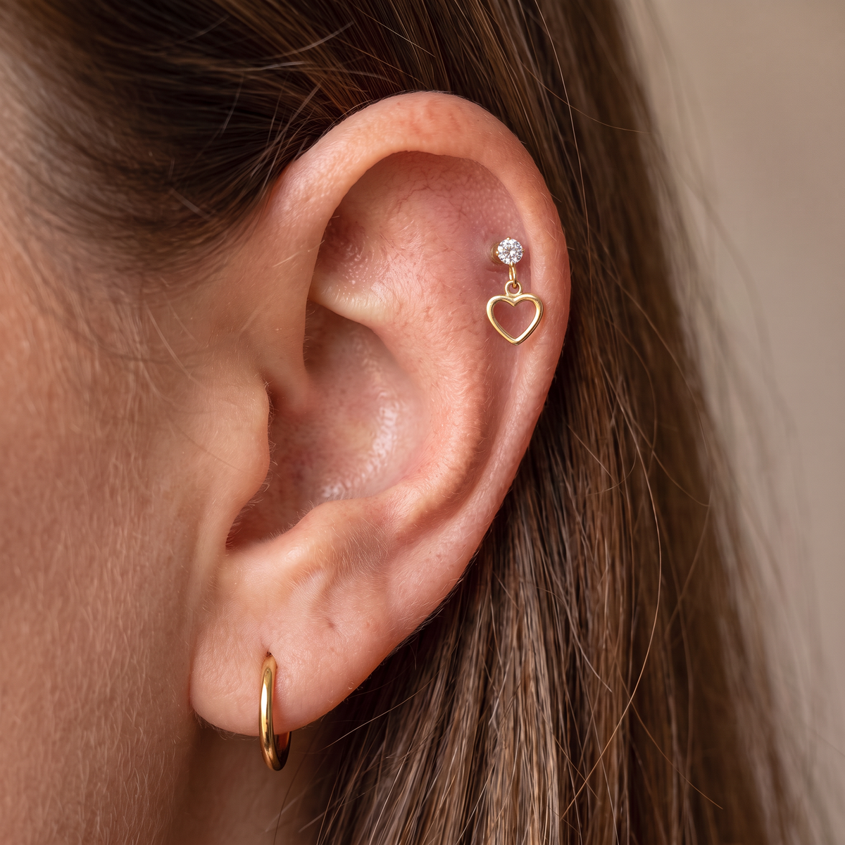 Close-up of a person's ear showcasing a gold ear cuff with a dangling open heart and a small clear gemstone stud, alongside a gold hoop earring.
