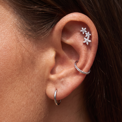 Close-up of a person's ear showcasing silver ear piercings: a small hoop in the lobe, a curved ear cuff with clear stones, and a cluster of three flower-shaped ear studs.