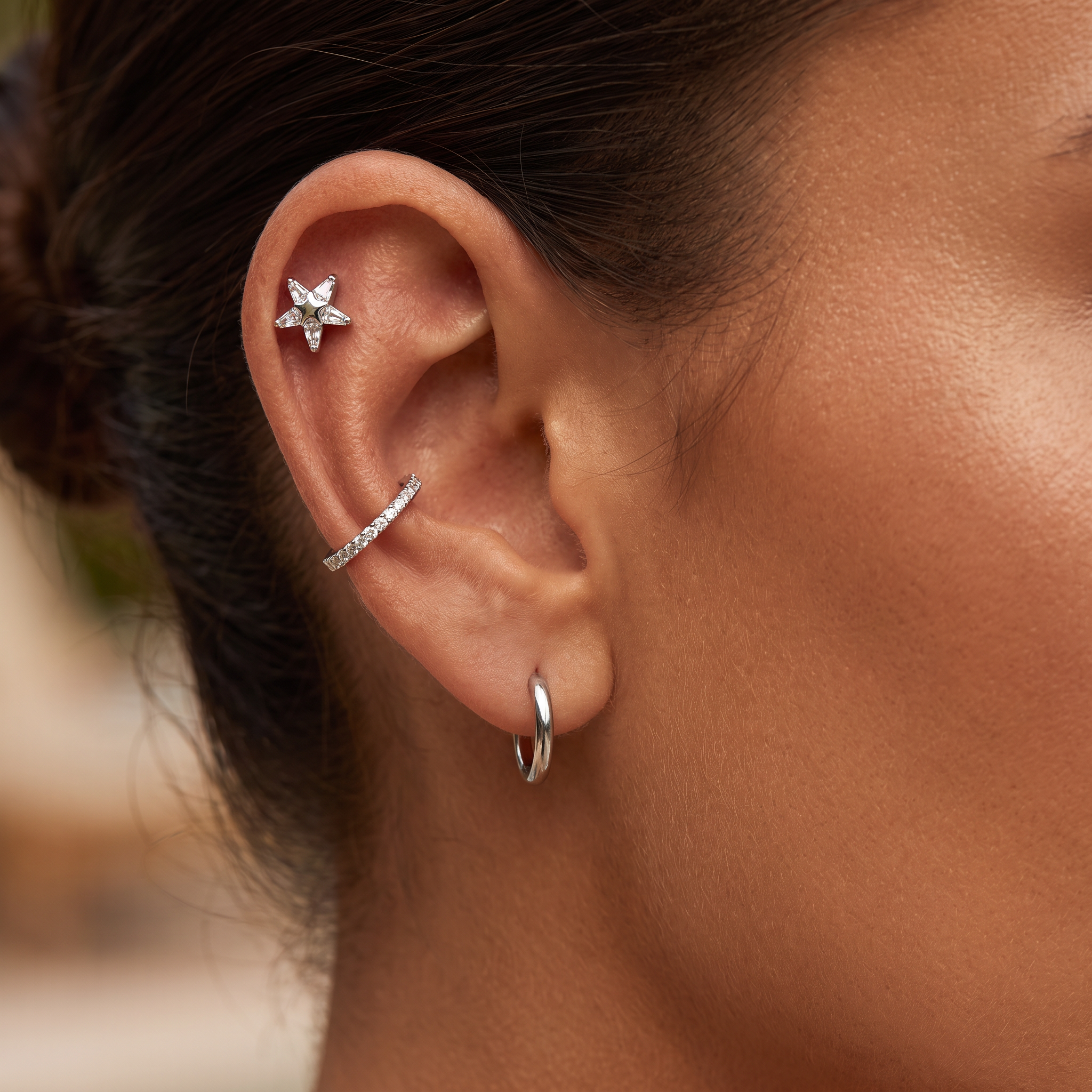 Close-up of a person's ear showcasing three silver ear piercings: a small star-shaped stud in the cartilage, a curved ear cuff with a pavé band, and a simple silver hoop in the lobe.