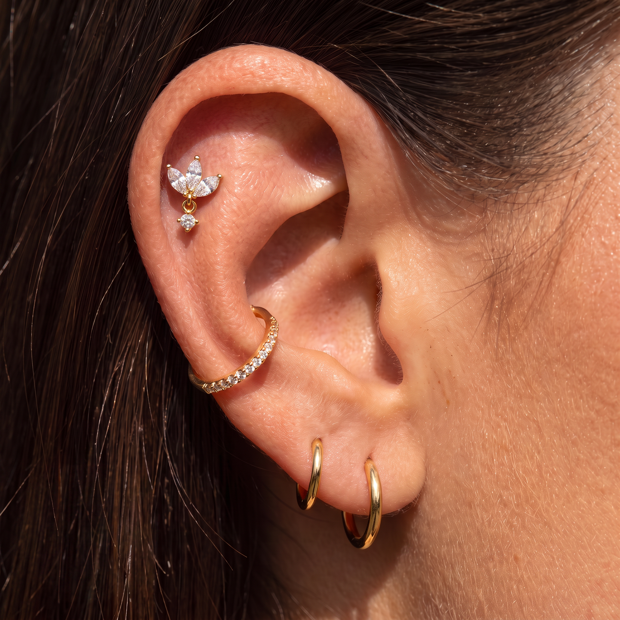 Close-up of a person's ear showcasing multiple gold and silver-toned ear piercings, including hoop earrings, a cartilage hoop, a butterfly-shaped cartilage stud with a dangling clear crystal, and a small lobe hoop.